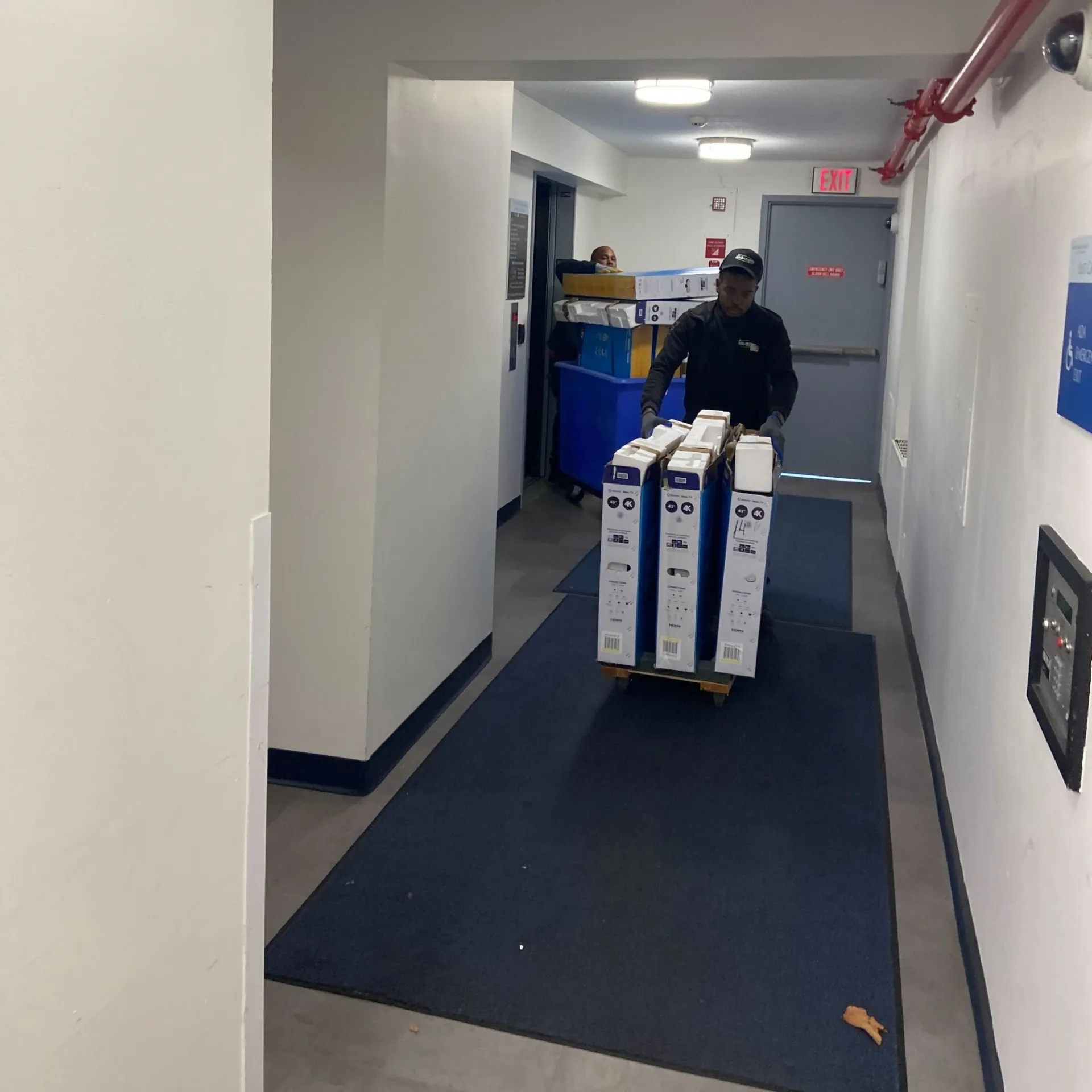 A staff member pushes a bin TVs on a cart through a carpeted hallway